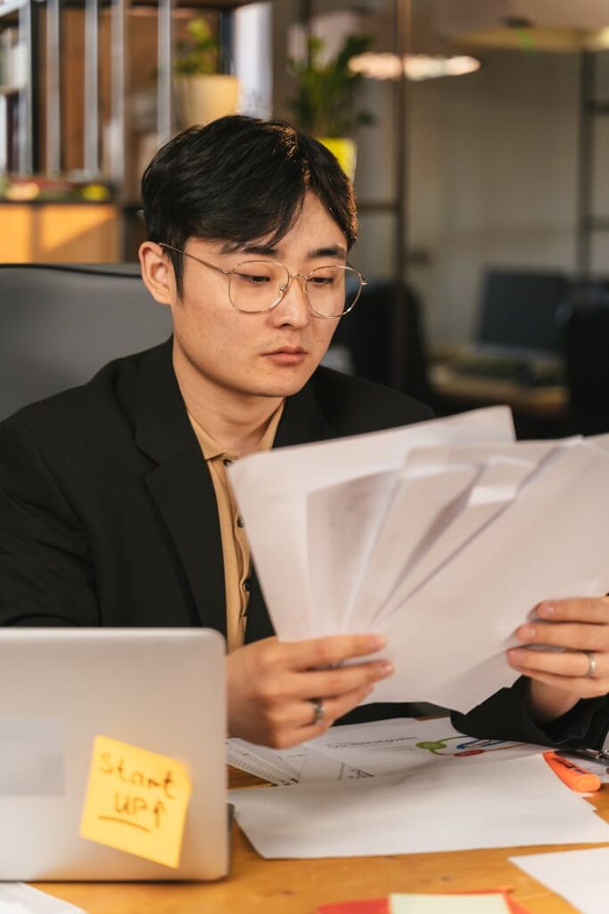Business professional in office meticulously reviews paperwork with laptop nearby.