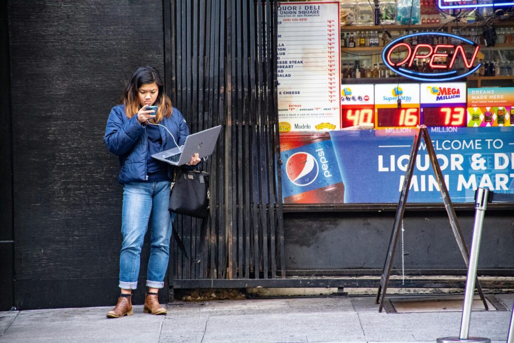 Asian woman using a laptop and phone outside a city store by an open sign.